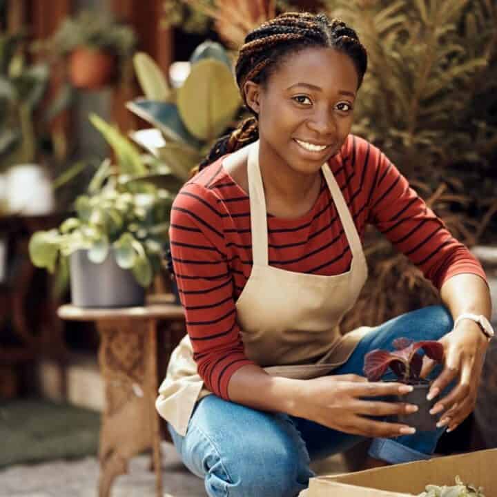 A woman wearing an apron and striped shirt smiles while potting a plant outdoors, surrounded by various potted plants and gardening supplies.