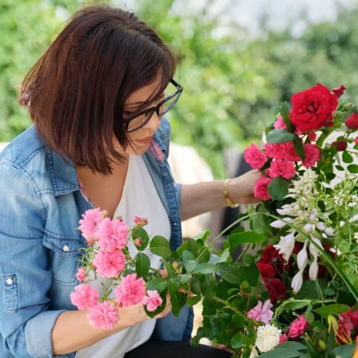 A woman in glasses and a denim shirt arranges a variety of colorful flowers outdoors.