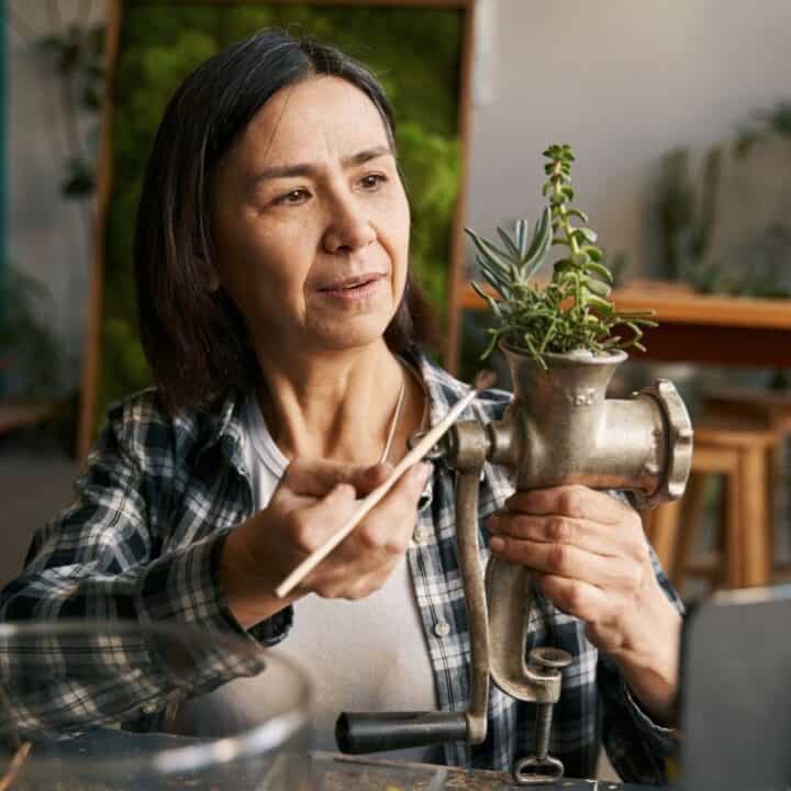 A woman trims a plant growing from an old metal grinder while sitting at a table in a well-lit room.