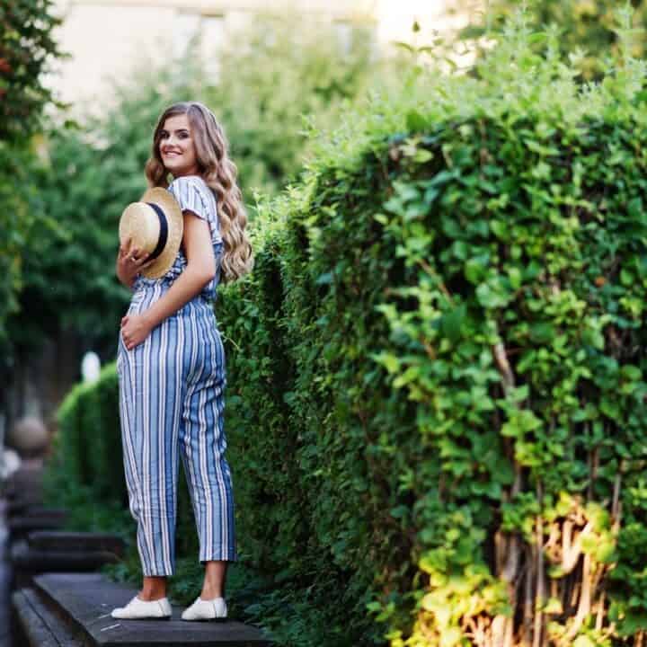 A woman in a blue and white striped jumpsuit stands on a path next to a green hedge, holding a straw hat and smiling.