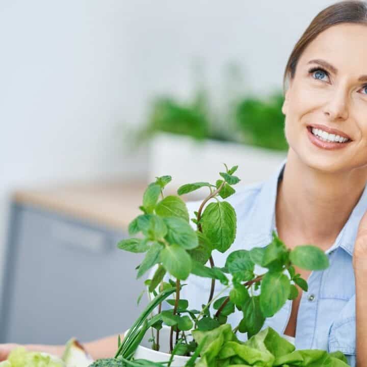 A woman holds a bottle of red juice next to her face and smiles, with fresh green herbs and vegetables in front of her on a kitchen counter.