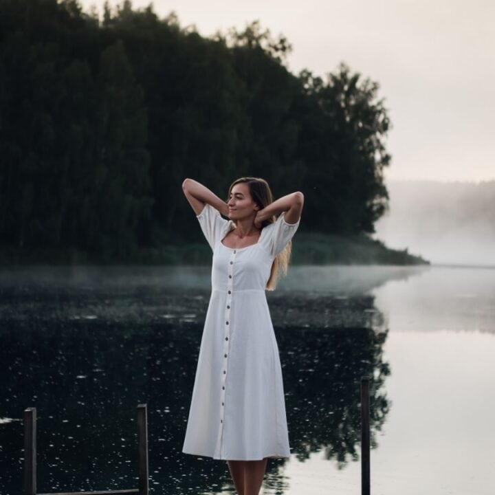 A woman in a white dress stands on a dock by a calm lake, with trees and mist in the background under a cloudy sky.