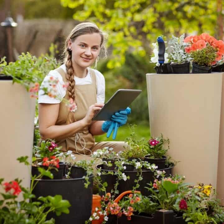 A woman in an apron and gloves is sitting among potted flowers outdoors, holding a tablet and smiling at the camera.