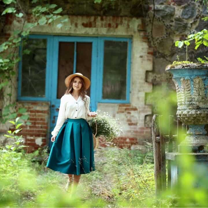 A woman in a hat and teal skirt stands in a garden holding flowers, in front of an old stone building with a blue door and large decorative planters.