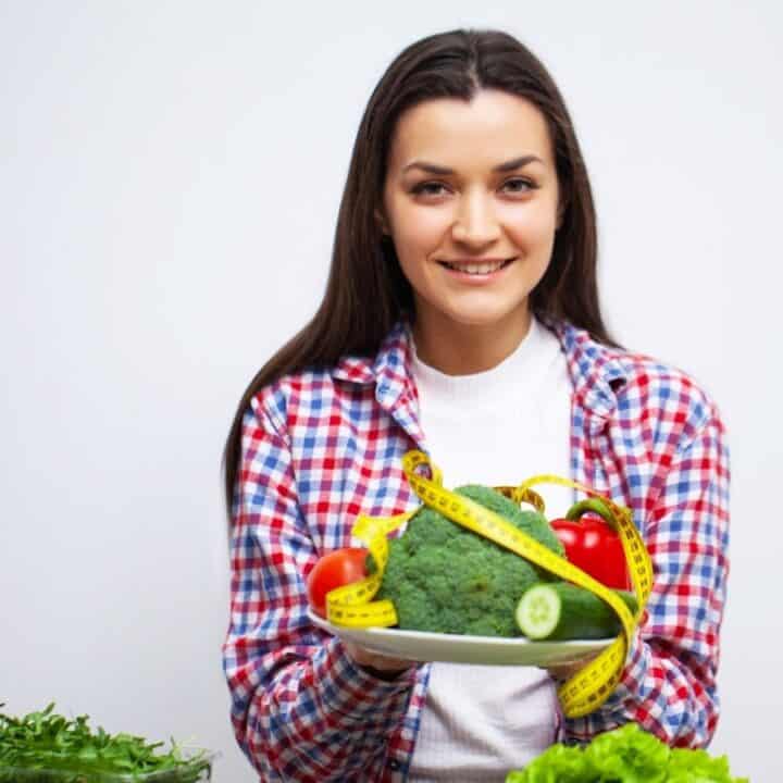 A woman in a plaid shirt holds a plate of vegetables, including broccoli and peppers, wrapped with a yellow measuring tape.