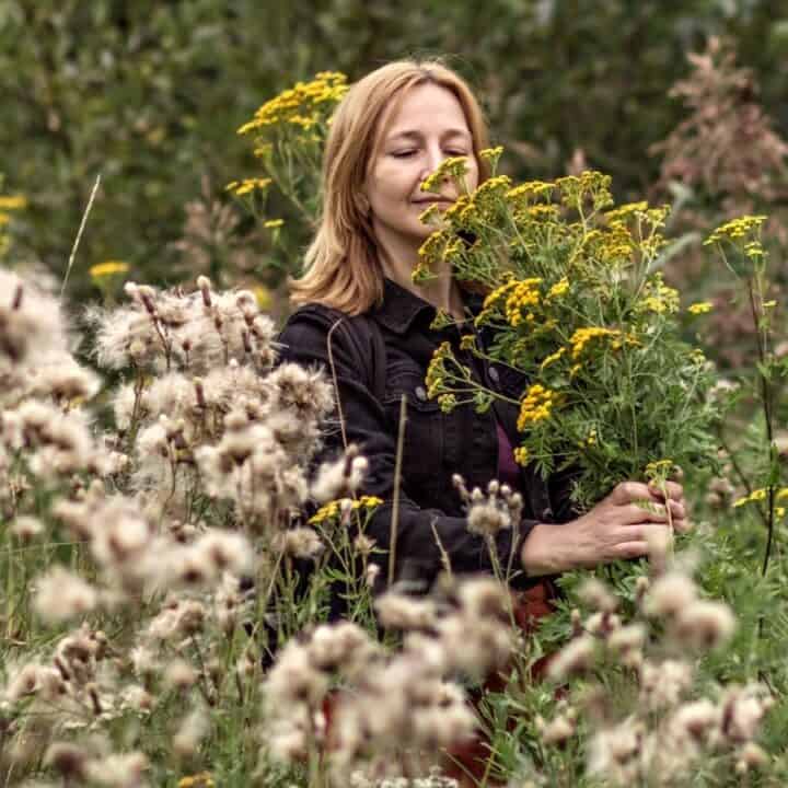 A woman stands in a field of wildflowers, holding a bouquet of yellow flowers and appearing to smell them.