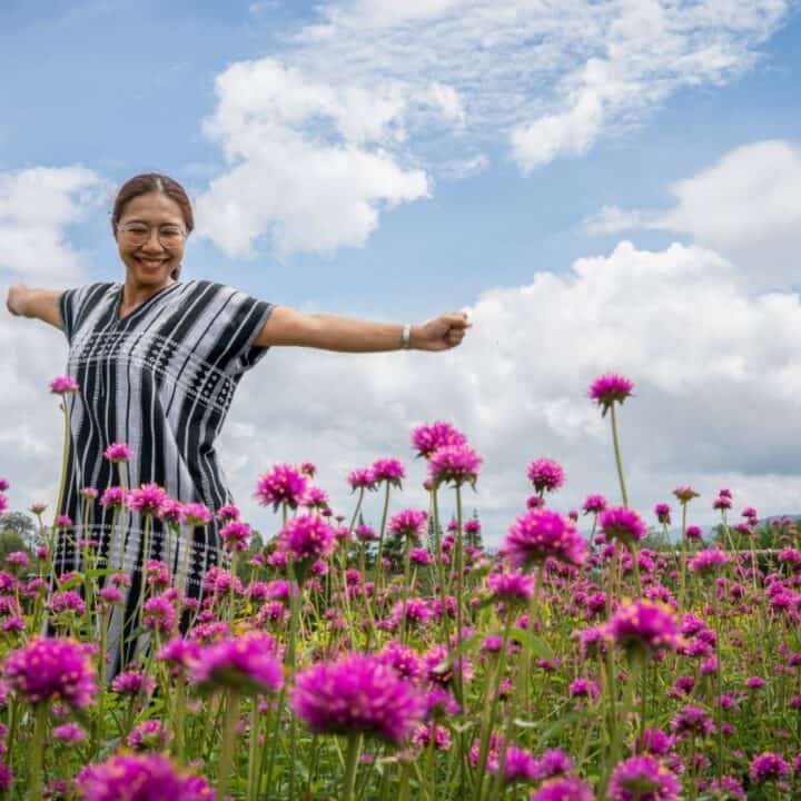 A person with outstretched arms stands in a field of pink flowers under a partly cloudy sky.