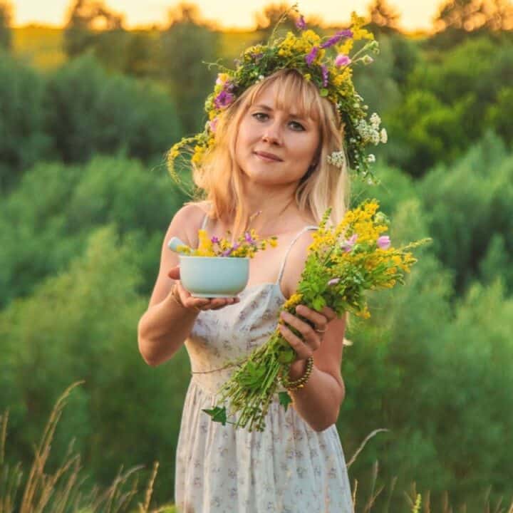 A woman in a floral dress stands outdoors holding a bouquet and a bowl of wildflowers, wearing a flower crown, with green trees in the background.