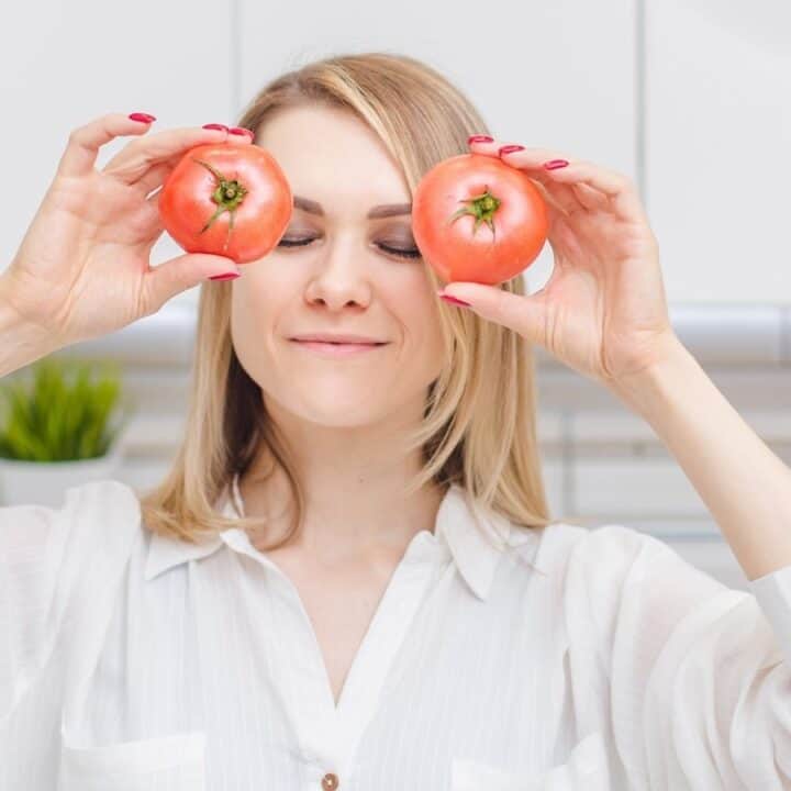 A woman stands in a kitchen holding two tomatoes over her eyes, smiling with her eyes closed.