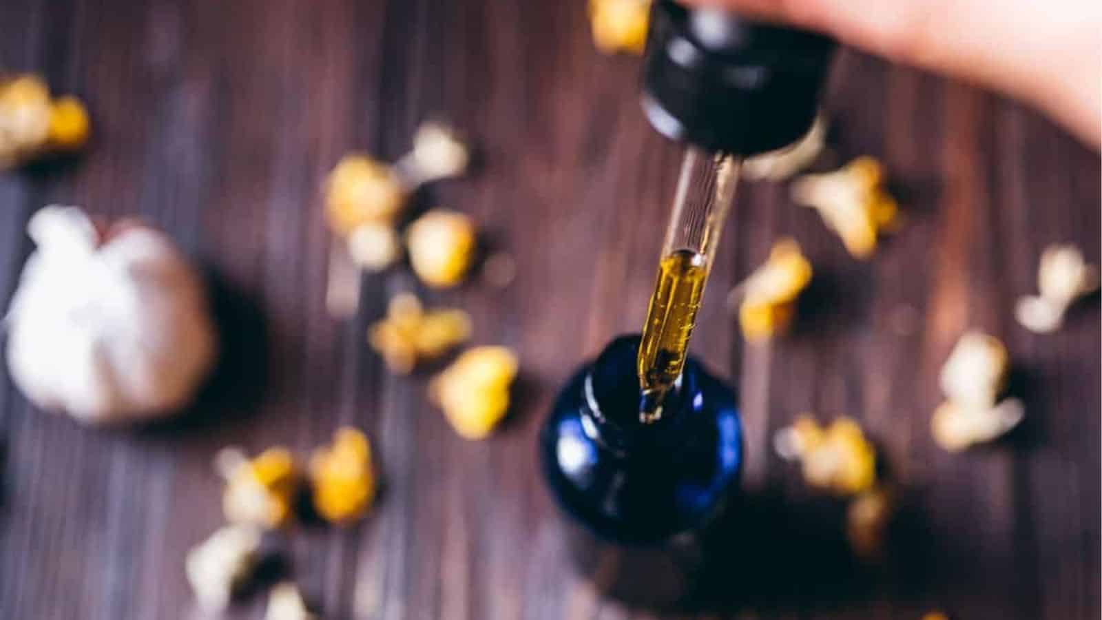 A hand holds a dropper above a dark glass bottle with yellow liquid, surrounded by dried flower pieces on a wooden surface.