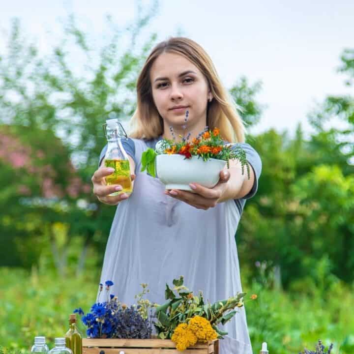 A woman standing outdoors holds a small bottle and a planter with flowers toward the camera. A table with flowers and greenery is in front of her. Trees and plants are in the background.