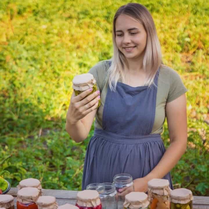 A woman wearing an apron stands outdoors, holding a jar of preserved food, with jars and fresh vegetables arranged on a table in front of her.