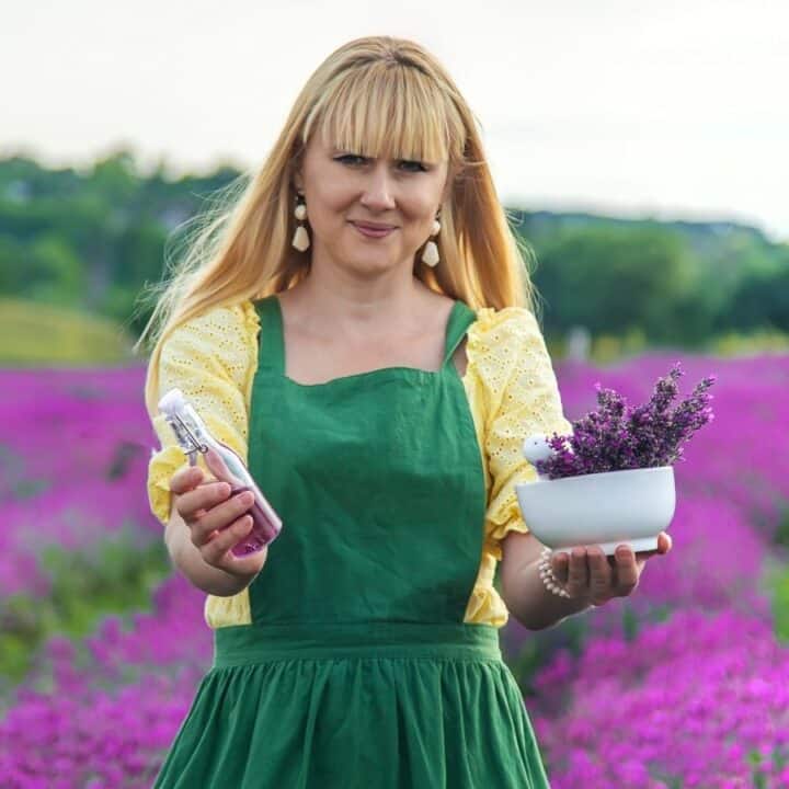 A woman in a green apron stands in a lavender field, holding a small bottle in one hand and a bowl of lavender flowers in the other.