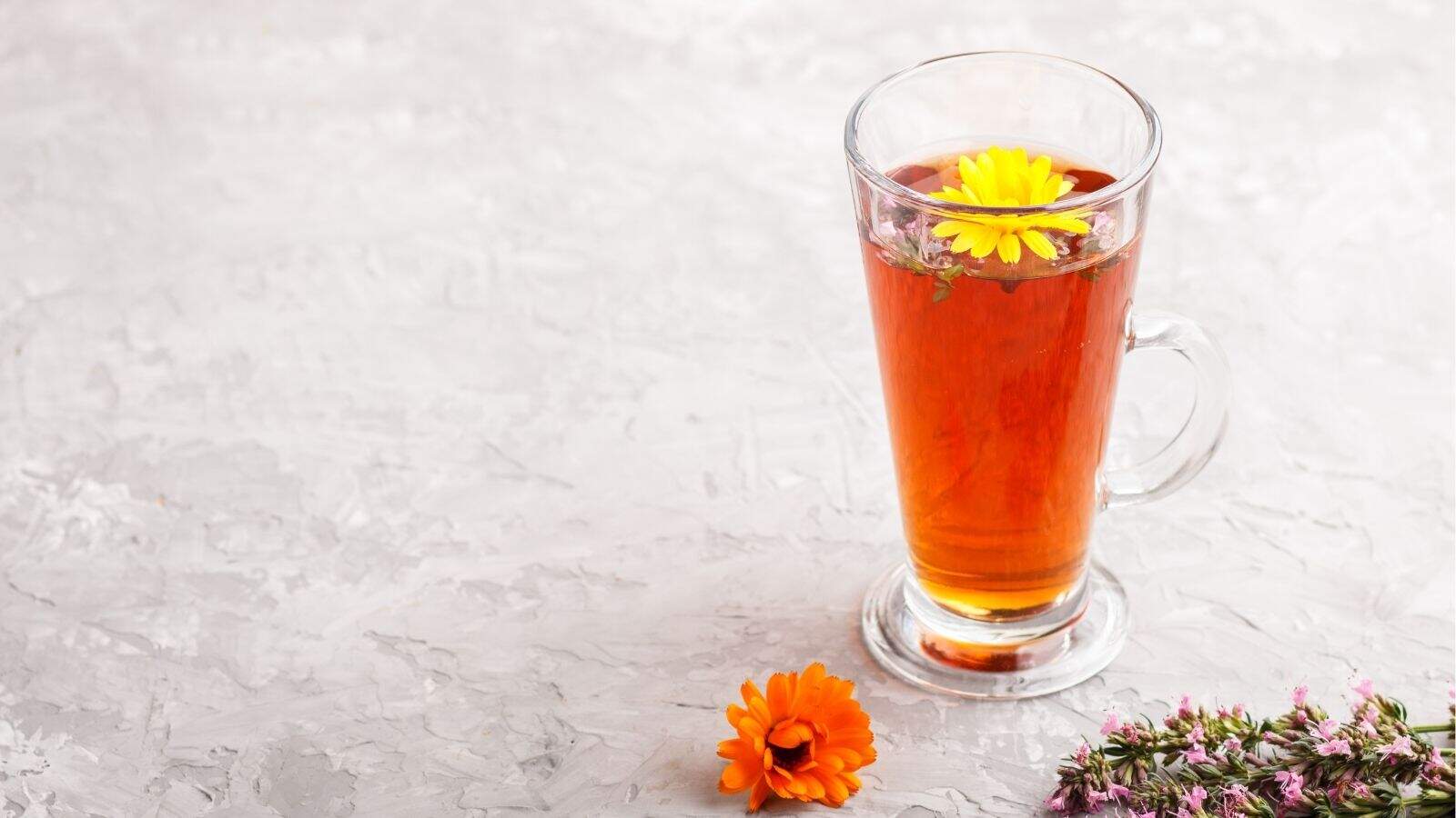 A glass mug of herbal tea with yellow flowers floating on top, placed on a light gray textured surface with more flowers nearby.