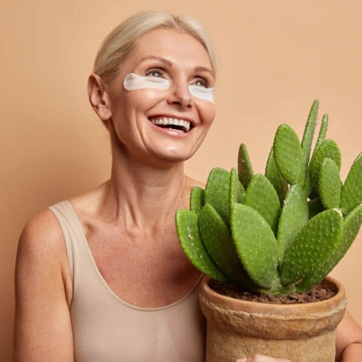 Smiling woman with under-eye patches holds a potted cactus against a plain beige background.
