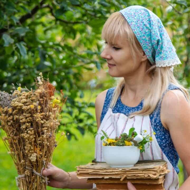 A woman outdoors holds a bundle of dried flowers in one hand and a stack of books with a bowl of fresh flowers in the other. She wears a floral headscarf and apron.