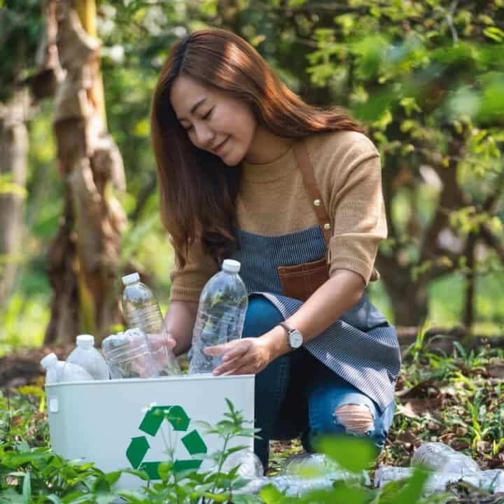A woman kneels outdoors, sorting plastic bottles into a white recycling bin surrounded by greenery.