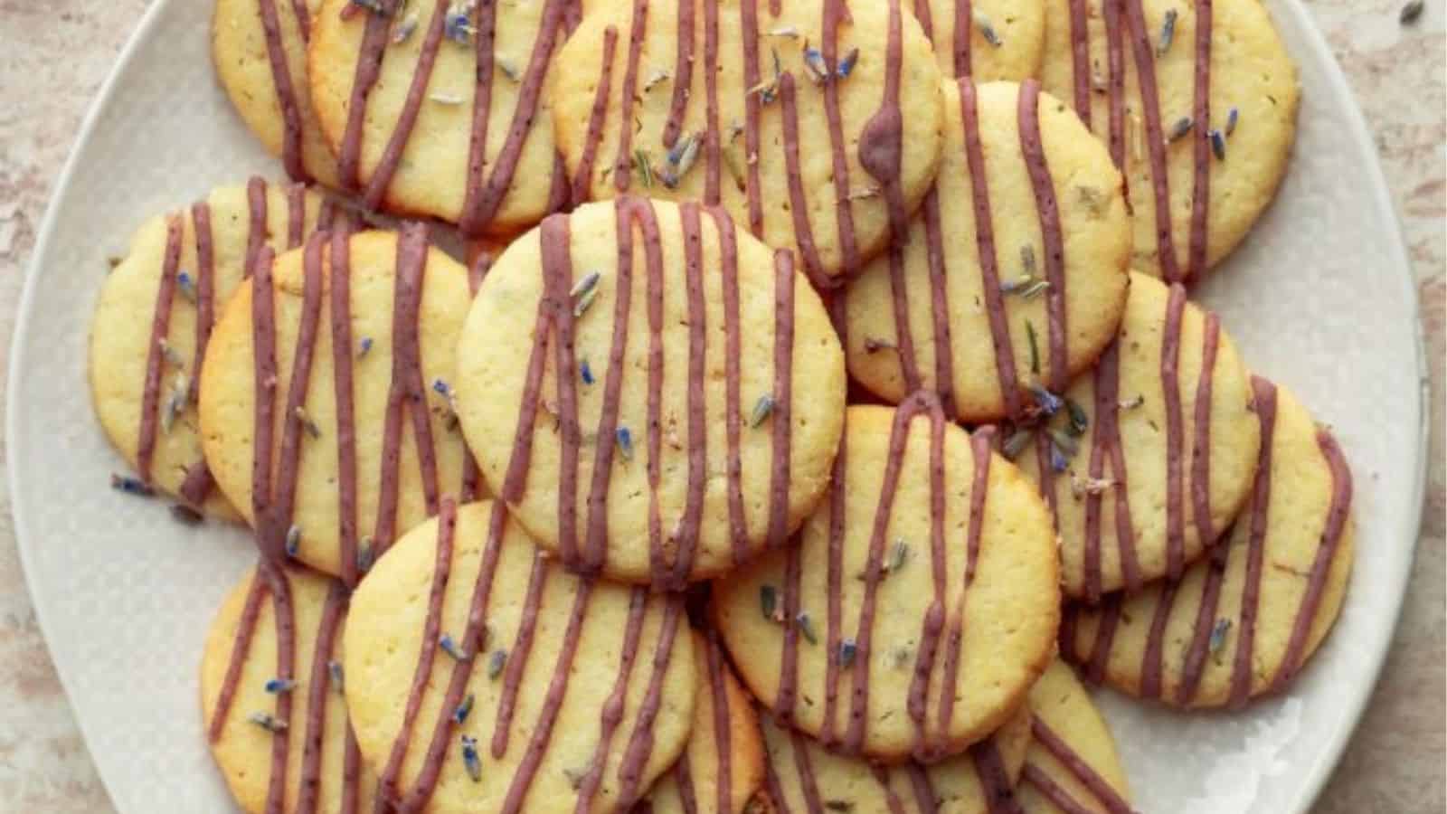 A plate of round, light-colored cookies drizzled with purple icing and sprinkled with small pieces of lavender.