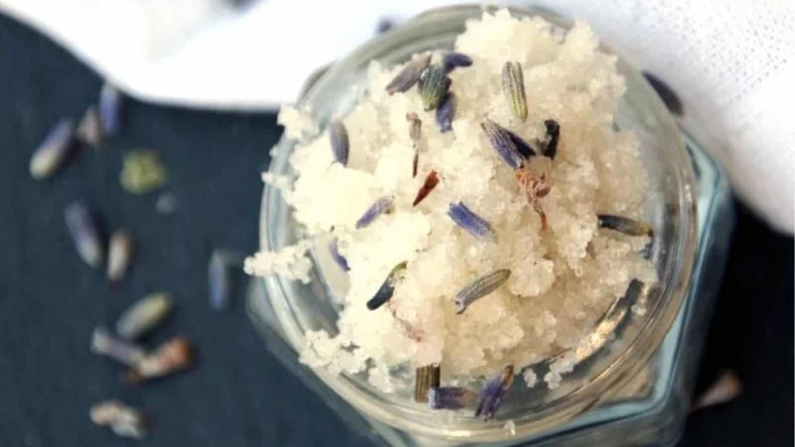 A glass jar filled with coarse white salt mixed with dried lavender buds, placed on a dark surface near a white cloth.