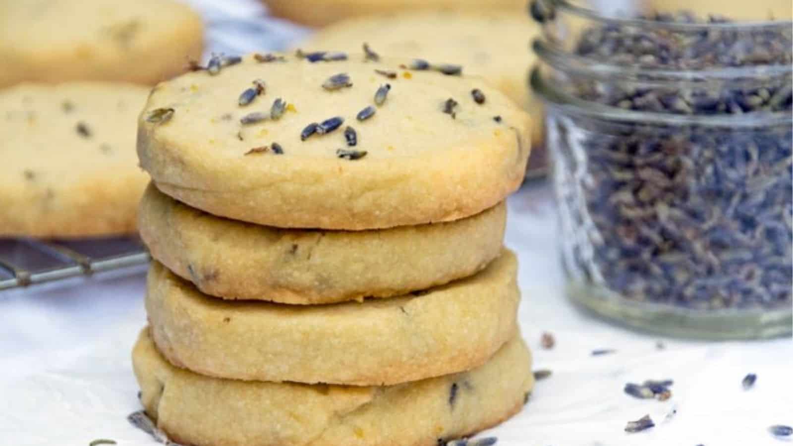 A stack of round lavender shortbread cookies with dried lavender sprinkled on top, next to a small jar of dried lavender buds.