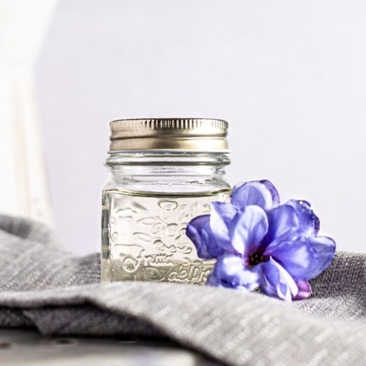 A small glass jar with a metal lid sits on a gray cloth next to a purple flower, with a white pitcher in the background.