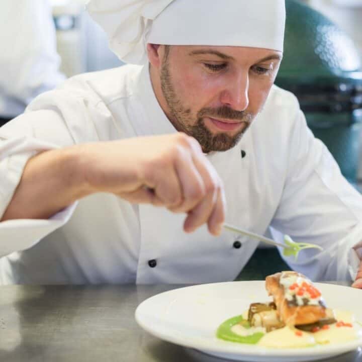 A chef in a white uniform and hat carefully garnishes a plated dish with herbs in a professional kitchen.