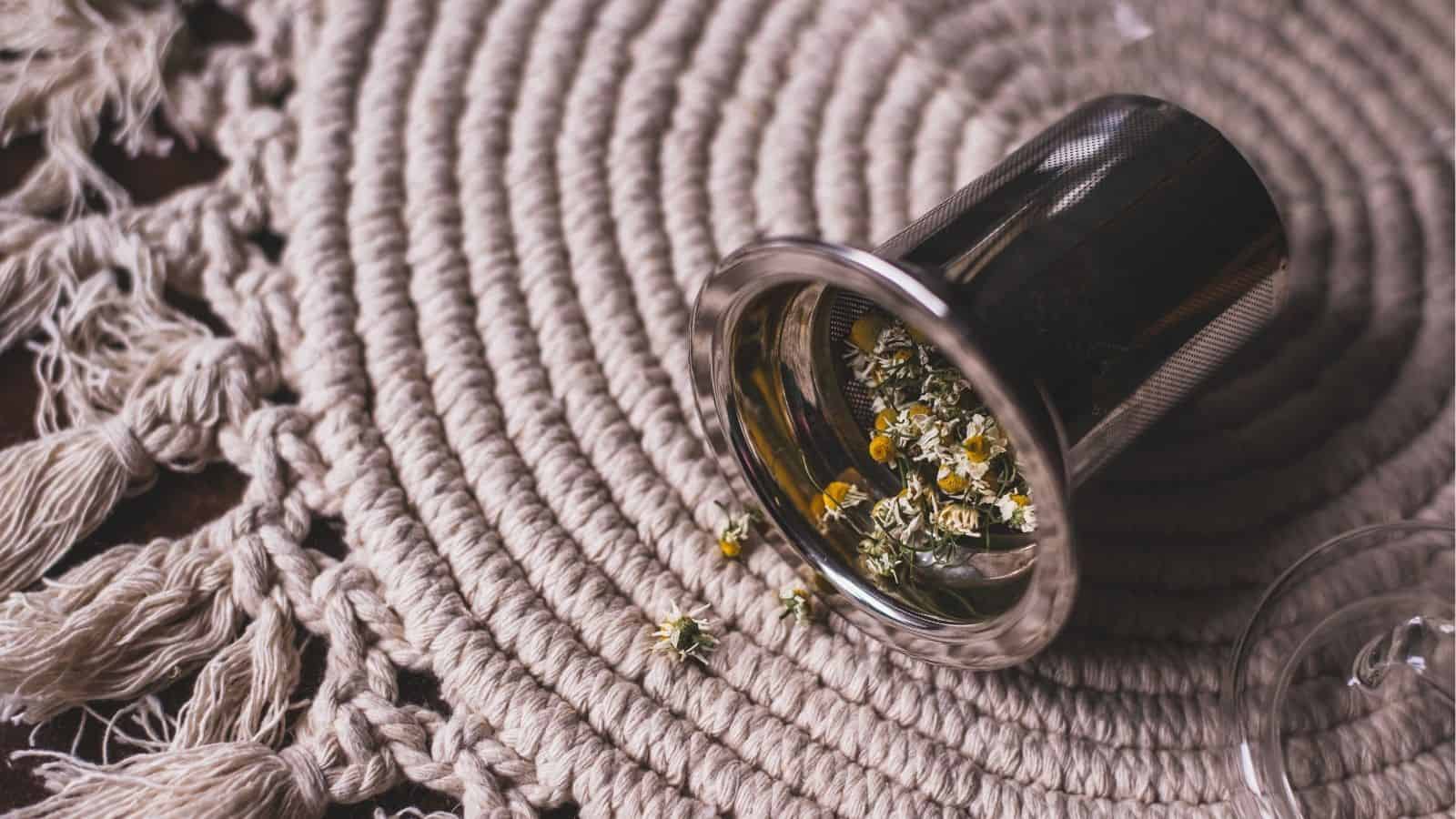 A metal tea infuser with loose dried chamomile flowers spilled on a woven, circular mat with fringed edges.