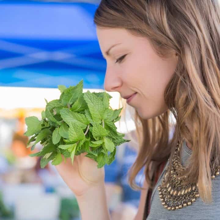 A woman holds and smells a bunch of fresh mint leaves at an outdoor market.