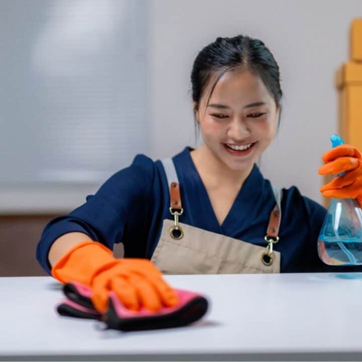 A woman wearing orange gloves and an apron cleans a white table with a spray bottle and cloth, smiling, with cardboard boxes stacked in the background.