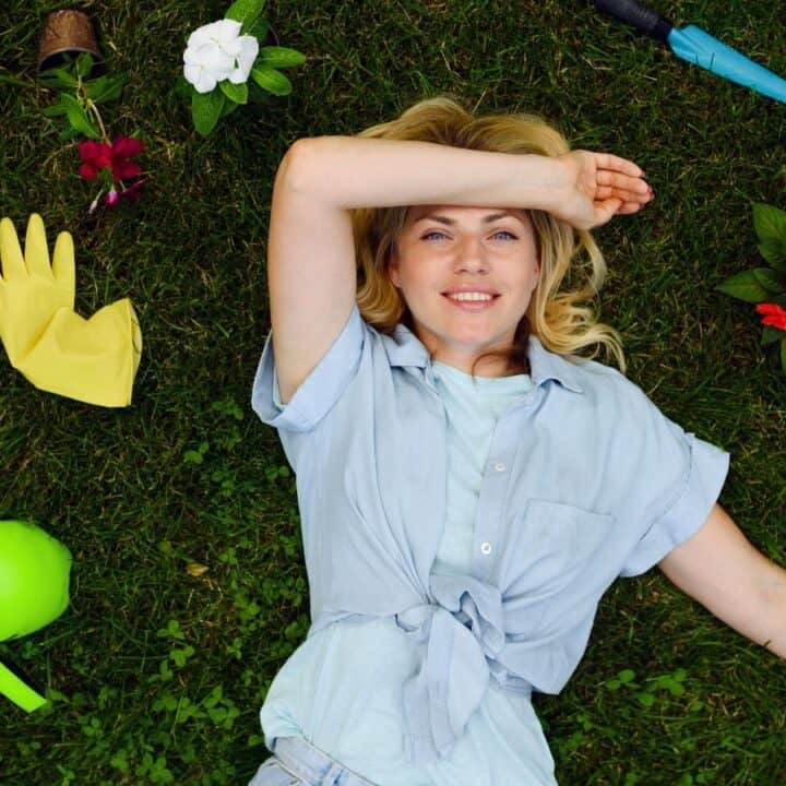 Woman lying on grass surrounded by gardening tools, gloves, spray bottle, and flowers, smiling and resting with one arm across her forehead.