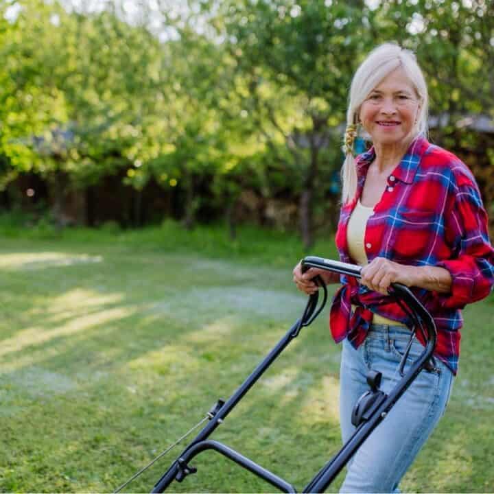 An older woman in a red plaid shirt and jeans is mowing the lawn with a push mower in a sunny, green backyard.
