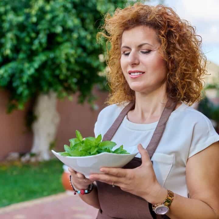 A woman with curly hair holds a bowl of fresh green herbs while standing outdoors in a garden area.