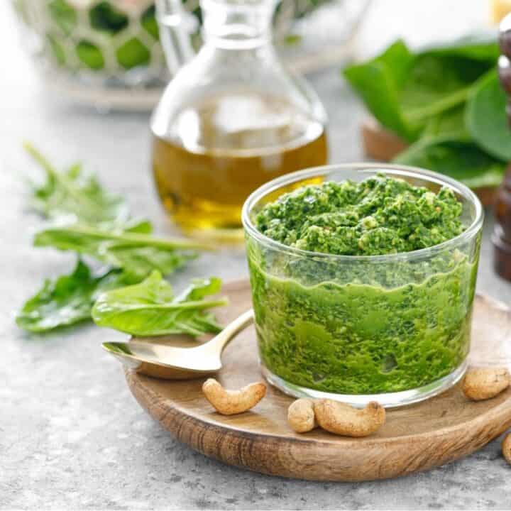A glass jar of green pesto sauce sits on a wooden plate with cashews, a gold spoon, olive oil, fresh basil, and salt and pepper shakers nearby.