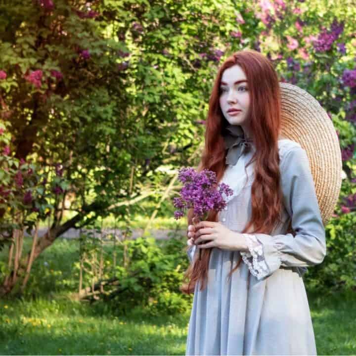 A young woman with long red hair in a light gray dress holds a lilac bouquet and stands outdoors by blooming bushes, wearing a wide-brimmed straw hat on her back.