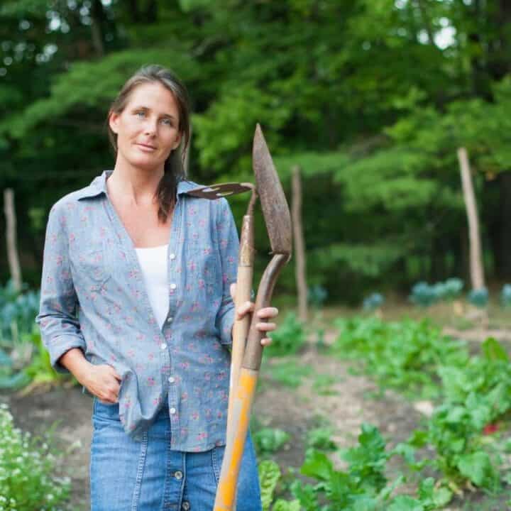 A woman stands in a vegetable garden holding a hoe, surrounded by green plants and trees in the background.