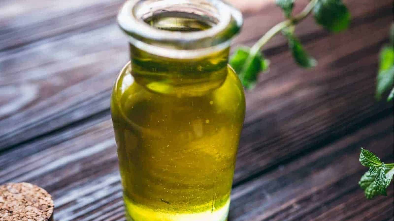 A glass bottle filled with light green oil sits on a wooden surface, with some green leaves and a cork nearby.