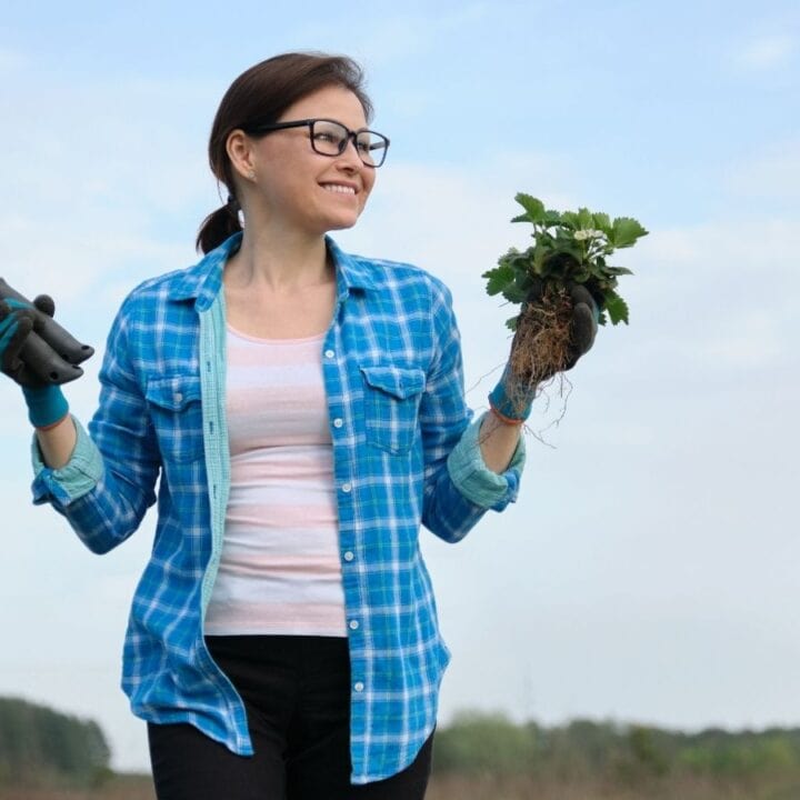 A woman wearing glasses and gloves holds a gardening trowel in one hand and a small plant with roots in the other, standing outdoors with trees in the background.