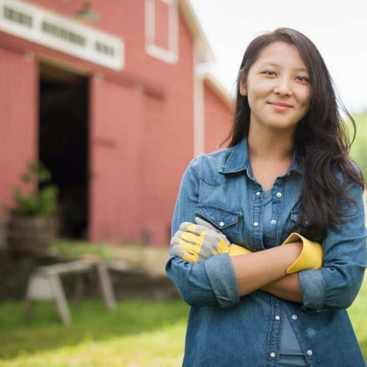 A woman wearing a denim shirt and yellow work gloves stands outside with arms crossed in front of a red barn on a sunny day.