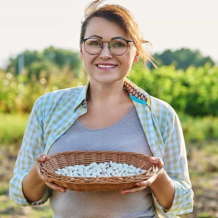 Woman standing outdoors in a field, holding a wicker basket filled with white beans, and smiling at the camera.