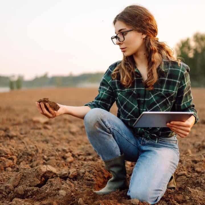 A person kneels in a field, examining soil with one hand and holding a tablet in the other, with trees in the background.