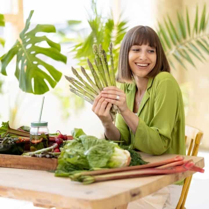 A woman sits at a table with various fresh vegetables, holding a bunch of asparagus and smiling. Large green plants are in the background.