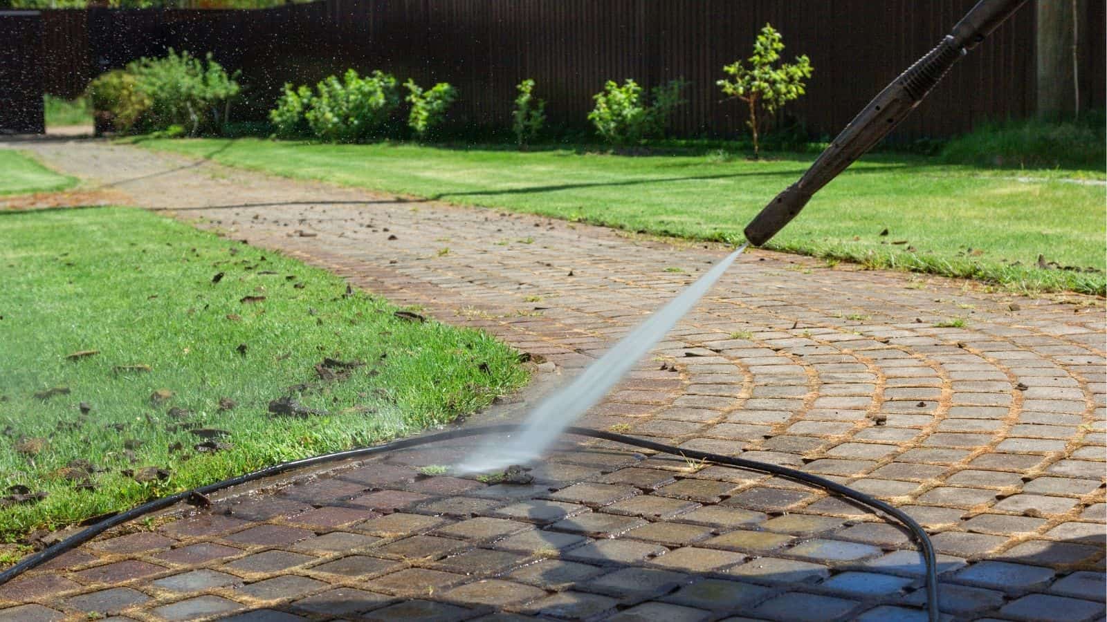 A person uses a pressure washer to clean a brick walkway in a garden with green grass and plants in the background.