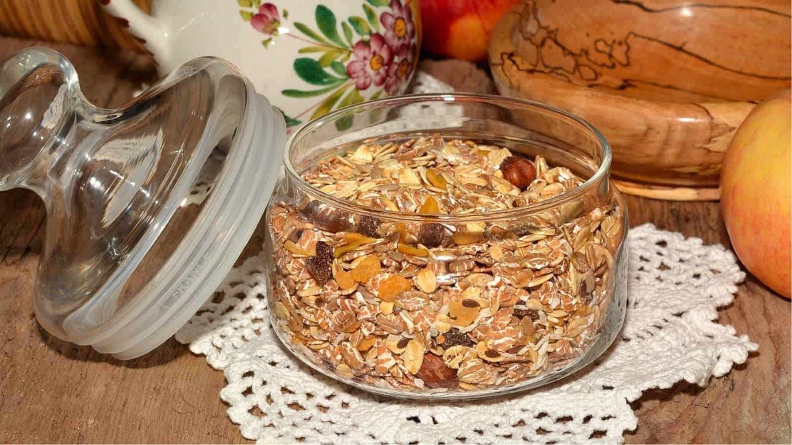 A glass jar filled with muesli sits on a crocheted doily on a wooden table, with a teapot, apple, and bowl in the background.