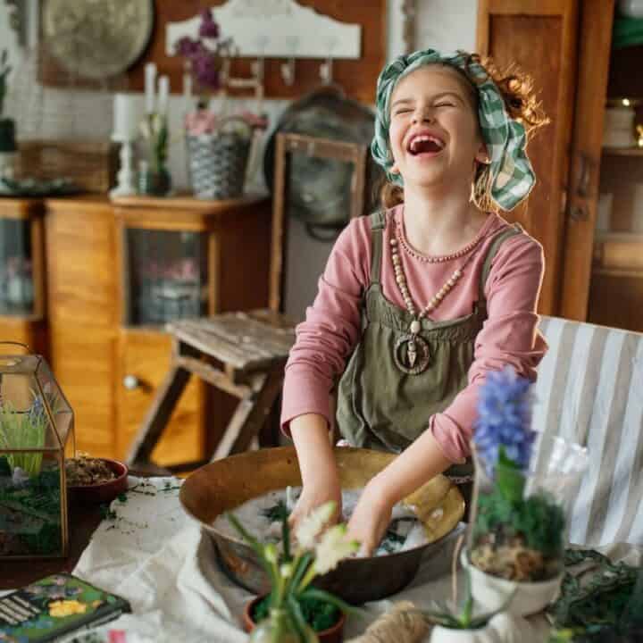 Young girl wearing a headscarf and apron laughs while mixing soil in a large bowl, surrounded by plants and gardening supplies on a table indoors.
