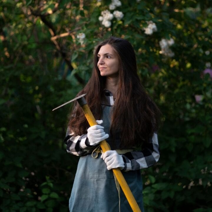 A woman in a checkered shirt and apron stands in a garden holding a yellow-handled hoe, wearing white gloves, with green foliage and white flowers in the background.