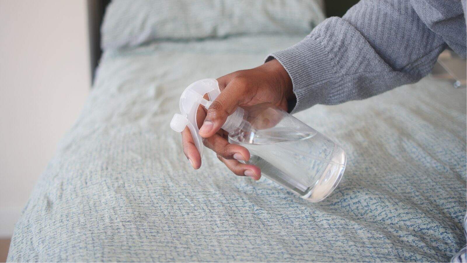 A person sprays liquid from a clear spray bottle onto a bed with light blue patterned bedding.