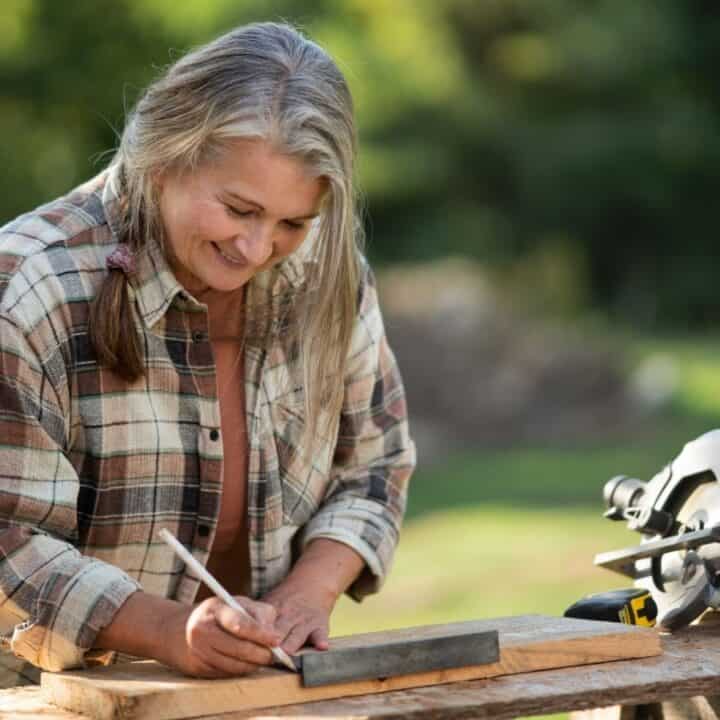 A woman in a plaid shirt measures and marks a piece of wood at an outdoor workbench, with a circular saw and other tools nearby.