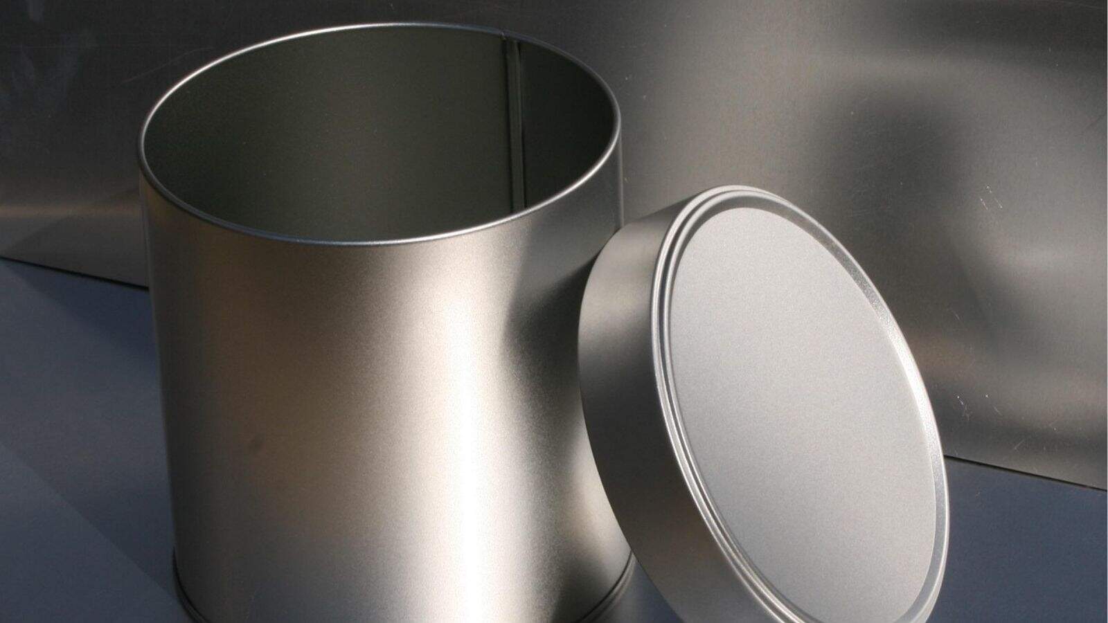 A large, empty, silver metal tin with its round lid removed and resting on its side next to the container.