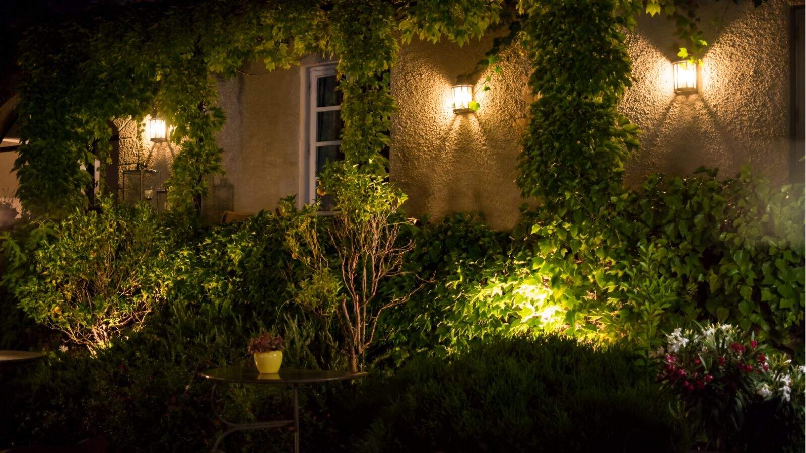 Outdoor wall with two lit lanterns, covered in lush green ivy and plants at night, with a window in the center and a small table holding a yellow pot in the foreground.