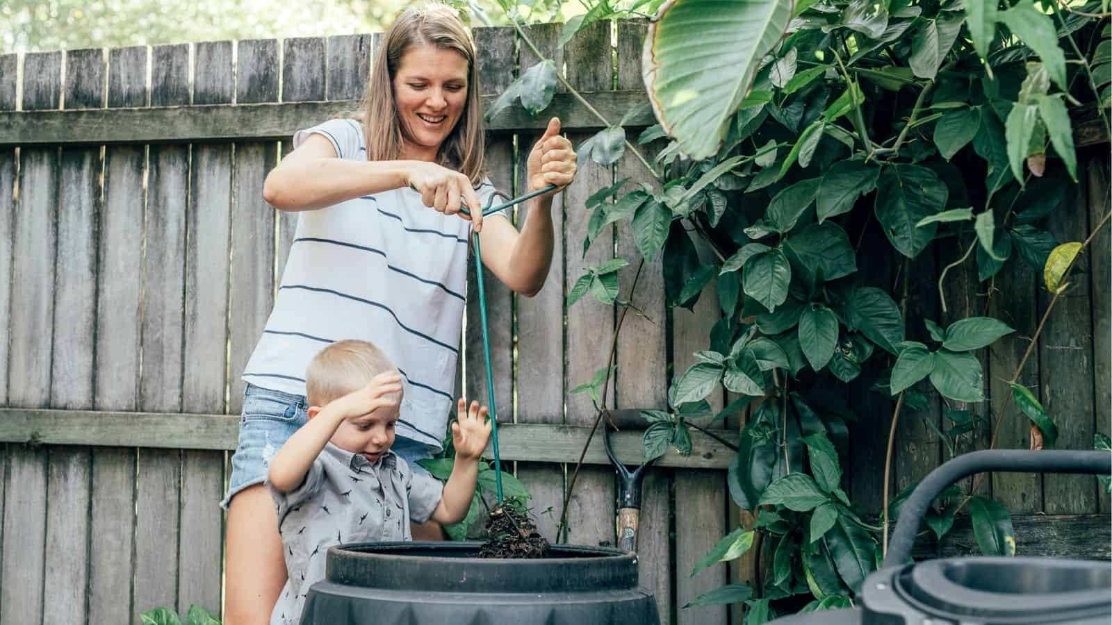 An adult and a child standing by a compost bin, stirring compost with a stick in a backyard garden next to a wooden fence and green plants.